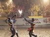Two knights in armor clash swords, causing sparks to fly, during a staged combat performance in an arena with an audience in the background.