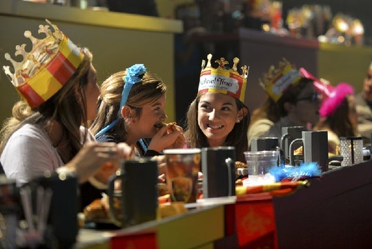 A group of people wearing paper crowns sit at a long table eating food and talking, with cups and plates in front of them in a festive indoor setting.