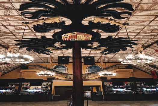 Large medieval-style hall with chandeliers, banners, a painted ceiling, and a central pillar featuring a decorative black crest with lions and shields.