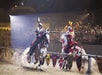 Two knights in armor joust on horseback in an arena, lances splintering on impact, while an audience watches from stands in the background.