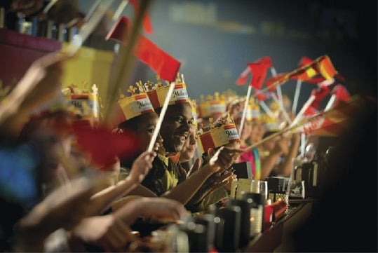 A group of people wearing paper crowns and waving flags sit at tables in a dimly lit arena, likely attending a themed dinner event.
