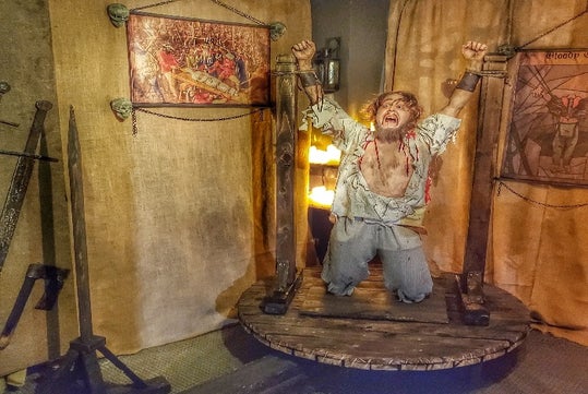 A mannequin depicting a tortured prisoner kneeling with wrists shackled, positioned in a medieval-style torture device exhibit.
