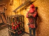 A person in a red hooded outfit stands beside a wooden cage containing another person, surrounded by medieval torture devices in a dimly lit room.