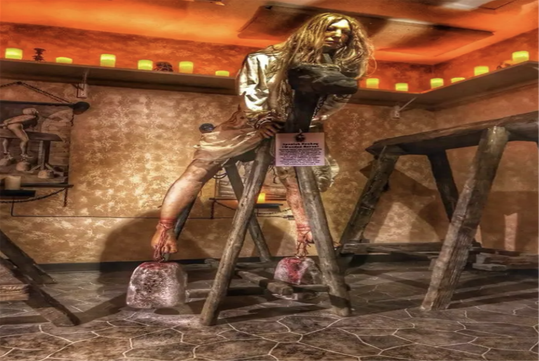 Museum display of a medieval torture device with a mannequin posed on a wooden frame, feet bound to weights, and a sign with exhibit information.