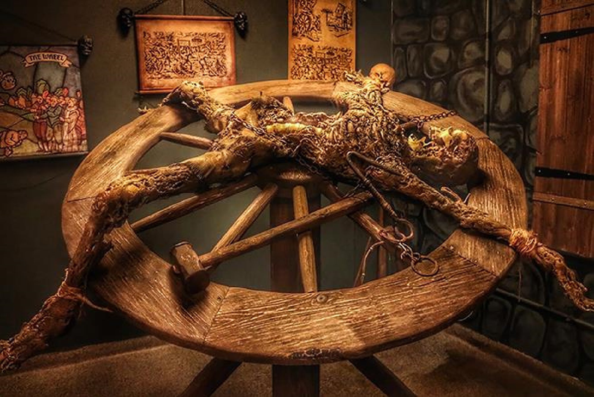A life-sized mannequin bound to a large wooden torture wheel, surrounded by medieval-themed decor and dim lighting.