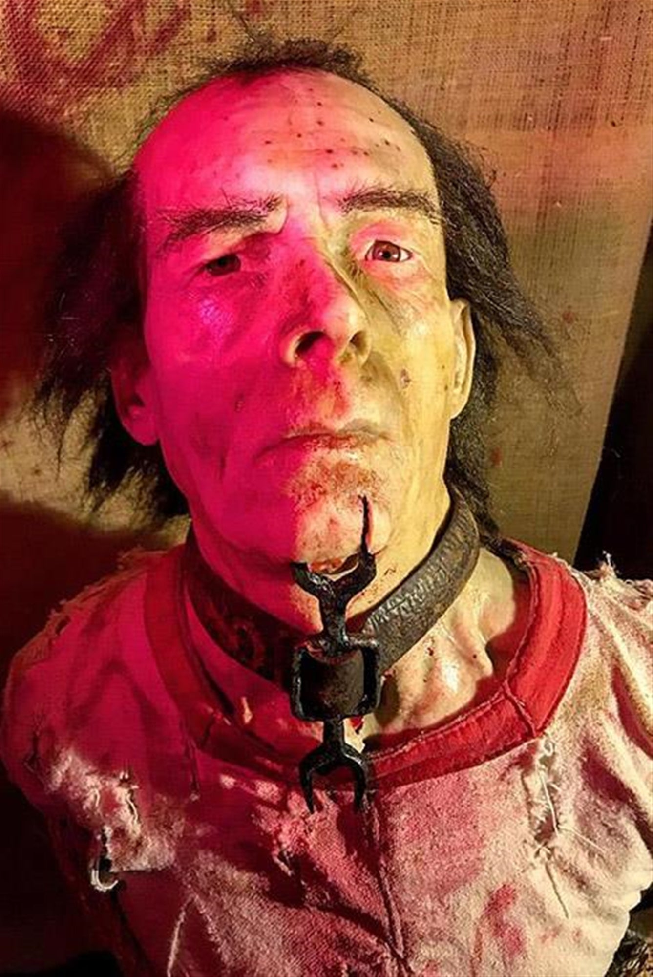 A mannequin resembling a disheveled man with a metal collar and clamp around his neck, wearing a torn, dirty shirt, lit with red lighting.
