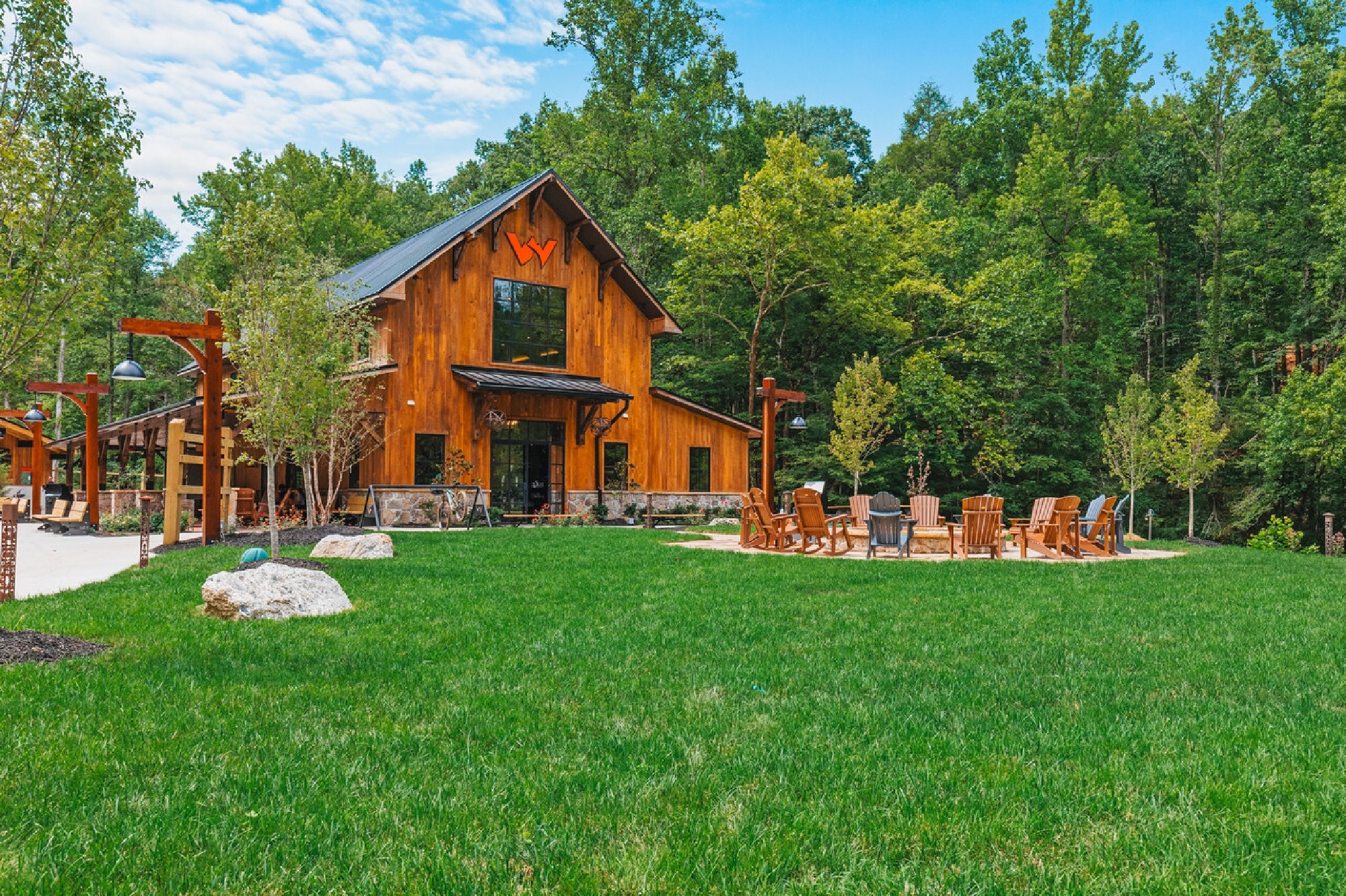 A wooden lodge with a large lawn, surrounded by trees, features outdoor seating around a firepit and a covered walkway on the left.