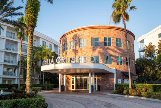 A modern hotel building with a round brick facade, large entrance canopy, and "AQUA ORLANDO" sign, surrounded by palm trees and greenery.