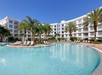 A large outdoor swimming pool with lounge chairs, palm trees, and a modern white multi-story hotel building in the background under a clear blue sky.