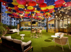 Outdoor bar lounge with string lights, colorful umbrellas suspended overhead, cushioned seating, and tables on artificial grass.