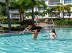 A man lifts a young child in the air while a woman watches at the edge of an outdoor pool surrounded by palm trees and a hotel building.