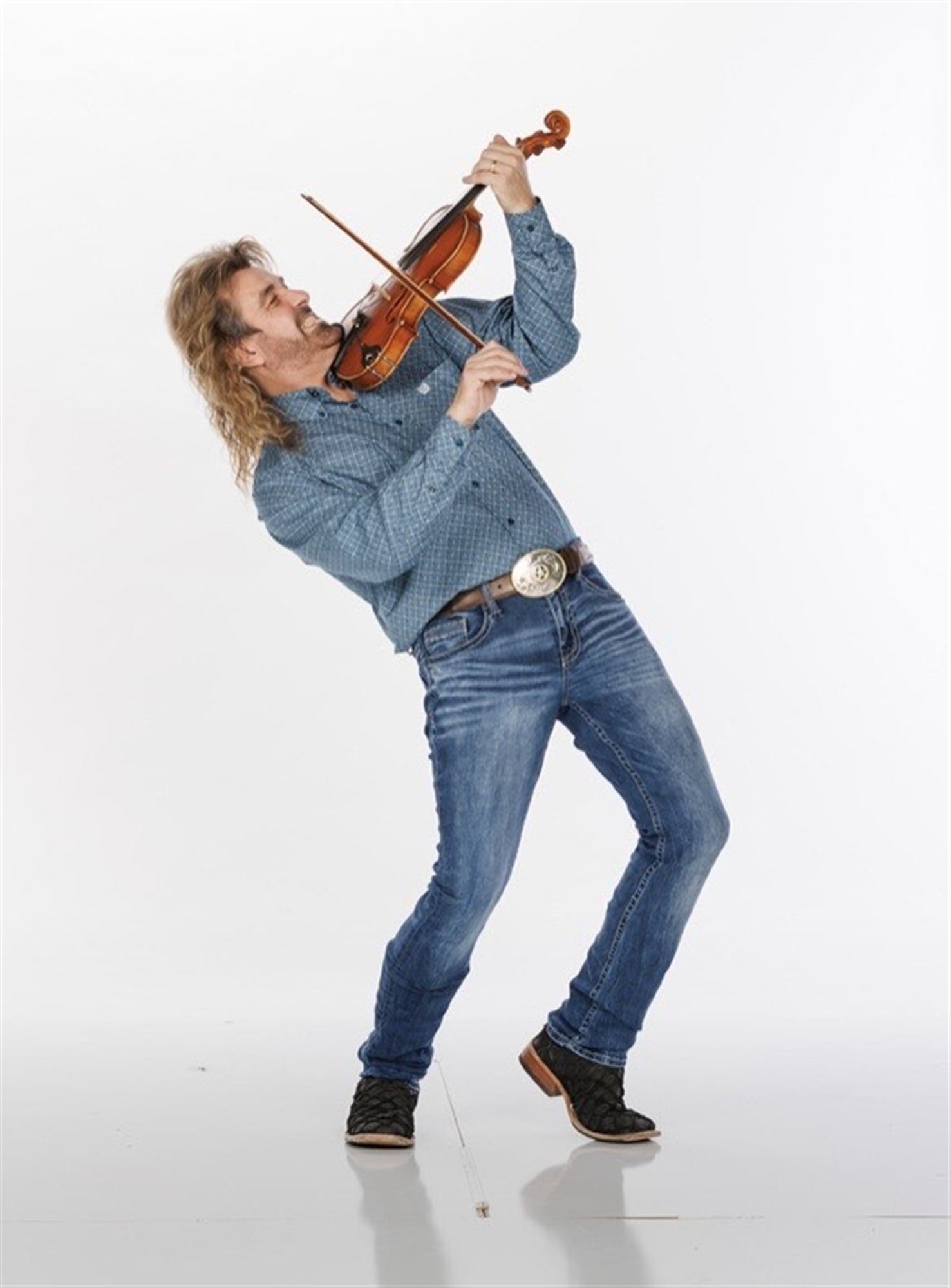 A man with long hair in a blue shirt and jeans stands and plays a violin energetically, leaning back with one foot forward against a plain white background.