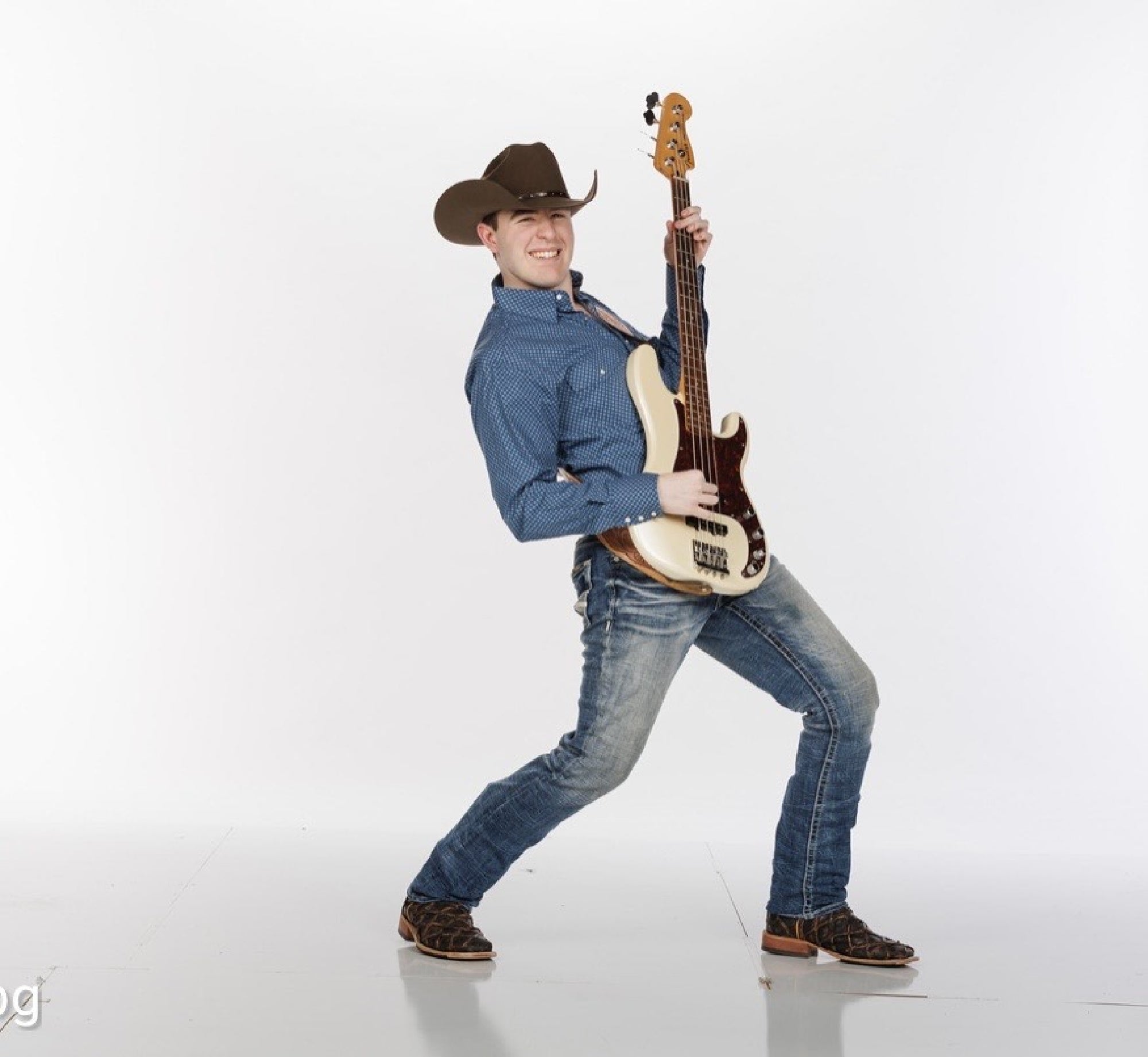 A man wearing a cowboy hat, blue shirt, and jeans plays an electric bass guitar while striking a wide, energetic pose against a plain white background.