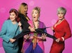 Four women pulling an oversized black bra in various directions with surprised and amused expressions against a pink background.