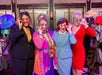 Four people striking playful poses and wearing colorful outfits and sunglasses, surrounded by colorful hats and accessories.