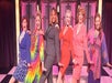 Six women are standing in a line on stage, dressed in colorful outfits, smiling, and posing with one leg raised.