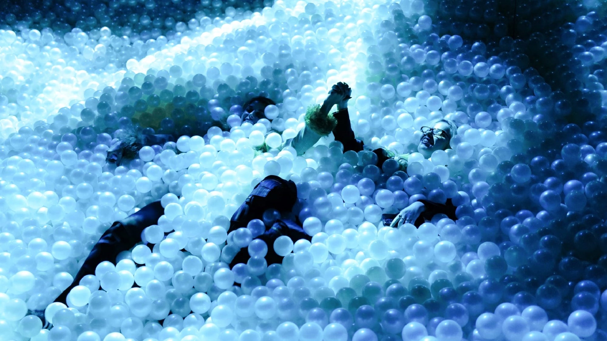 Two people partially submerged in a large pool filled with white plastic balls, holding hands and looking up under blue lighting.
