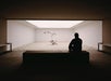 Silhouette of a person sitting on a bench, viewing a robotic arm in a minimalist, sand-filled room.
