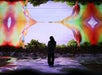 A person stands in front of a vibrant digital display featuring colorful abstract shapes and green trees, with reflections on the ground.