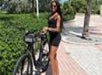 A woman in a black outfit stands beside a bicycle on a paved path surrounded by greenery.