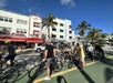 A group of cyclists stops on a sunny street with palm trees alongside art deco buildings, including a hotel, under a clear blue sky.