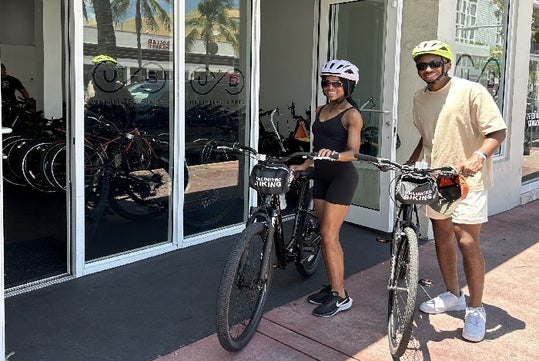 Two people with helmets stand outside a bike rental shop holding bicycles. Bikes are visible through the glass doors behind them.