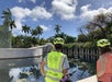Two people in helmets and safety vests stand by a reflective water feature with a large hand sculpture. Tropical trees and cloudy blue sky are in the background.