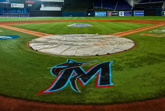 A view of a baseball field from behind home plate, featuring the Miami Marlins logo painted on the grass and a tarp covering the pitcher's mound.
