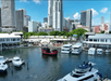 A harbor with several docked boats and a red ship in the water. Tall buildings and a shopping area are visible in the background under a clear blue sky.