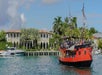 A red and black pirate ship sails by a luxurious waterfront mansion with palm trees and a cloudy sky.