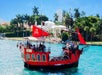 A red pirate-themed ship with black details and a skull and crossbones flag sails in calm blue water, with palm trees and buildings visible in the background.