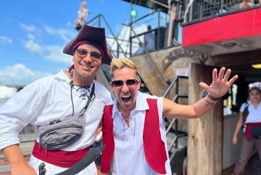 Two people in pirate-themed attire smiling at the camera, standing on a ship deck. One wears a tricorn hat and a gray sash; the other has sunglasses and raises a hand.