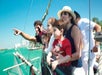 A group of people, including children, stand on a boat looking out at the water. One person points toward the distance. The sky is clear and the water is a bright turquoise.
