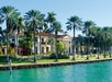 A luxurious waterfront mansion with palm trees, large windows, and a stone dock. The building features classical architectural elements, including columns, and overlooks a vibrant turquoise waterway.