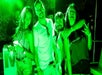 Four people smiling and posing under green lighting at a party. One person holds a drink, and another makes a peace sign.