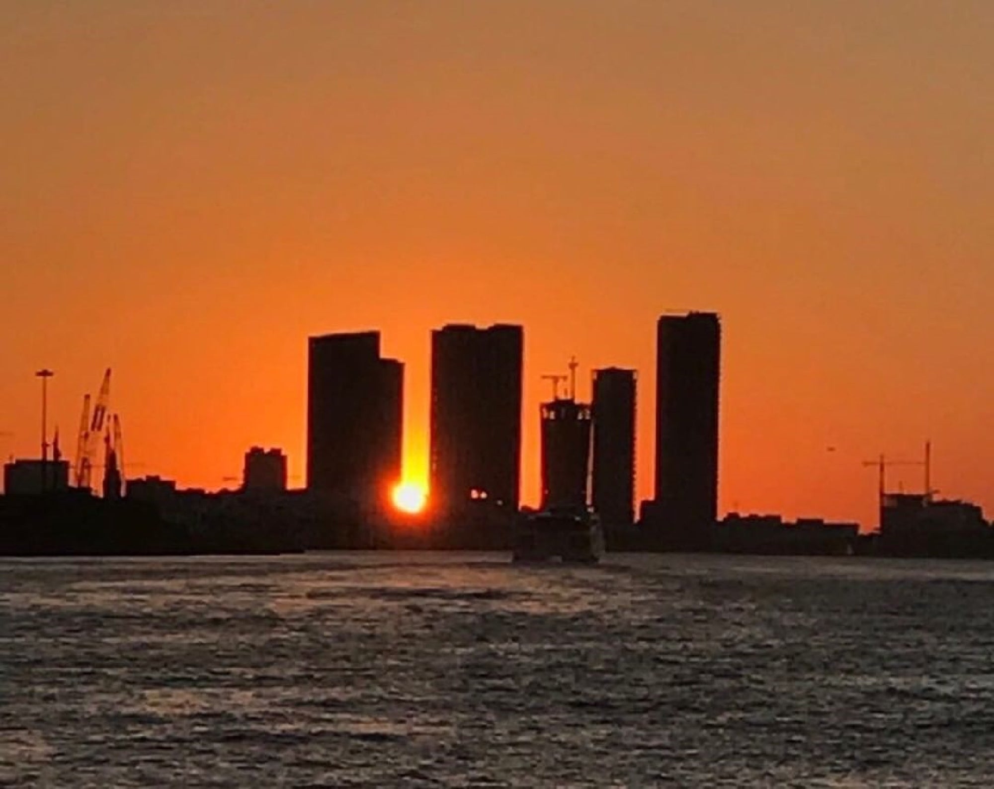 Sunset silhouette of a city skyline with tall buildings against an orange sky, a body of water in the foreground, and cranes visible on the left.