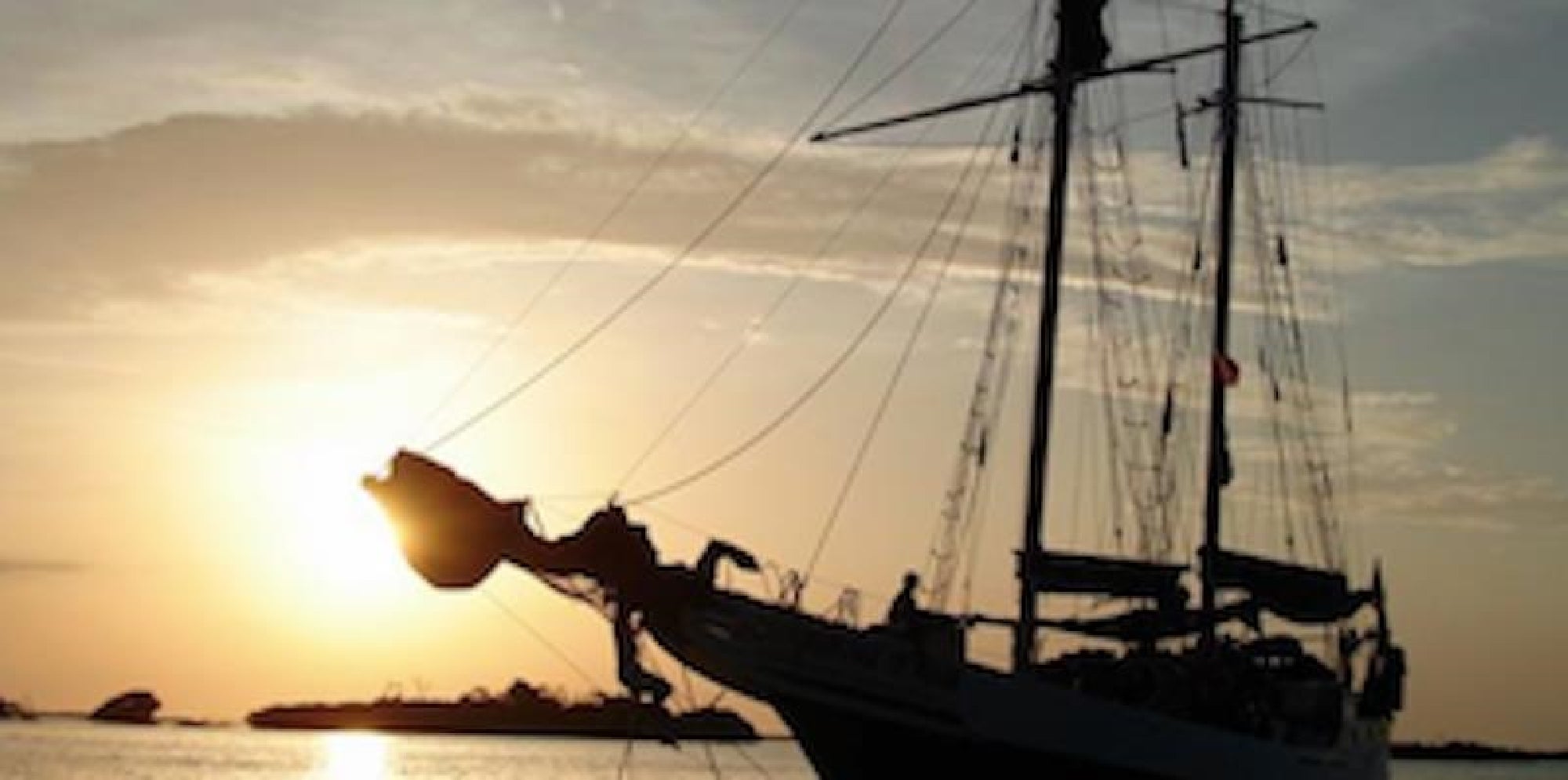 A sailing ship silhouetted against a sunset, with visible masts and rigging. The sun is partially obscured by the ship.