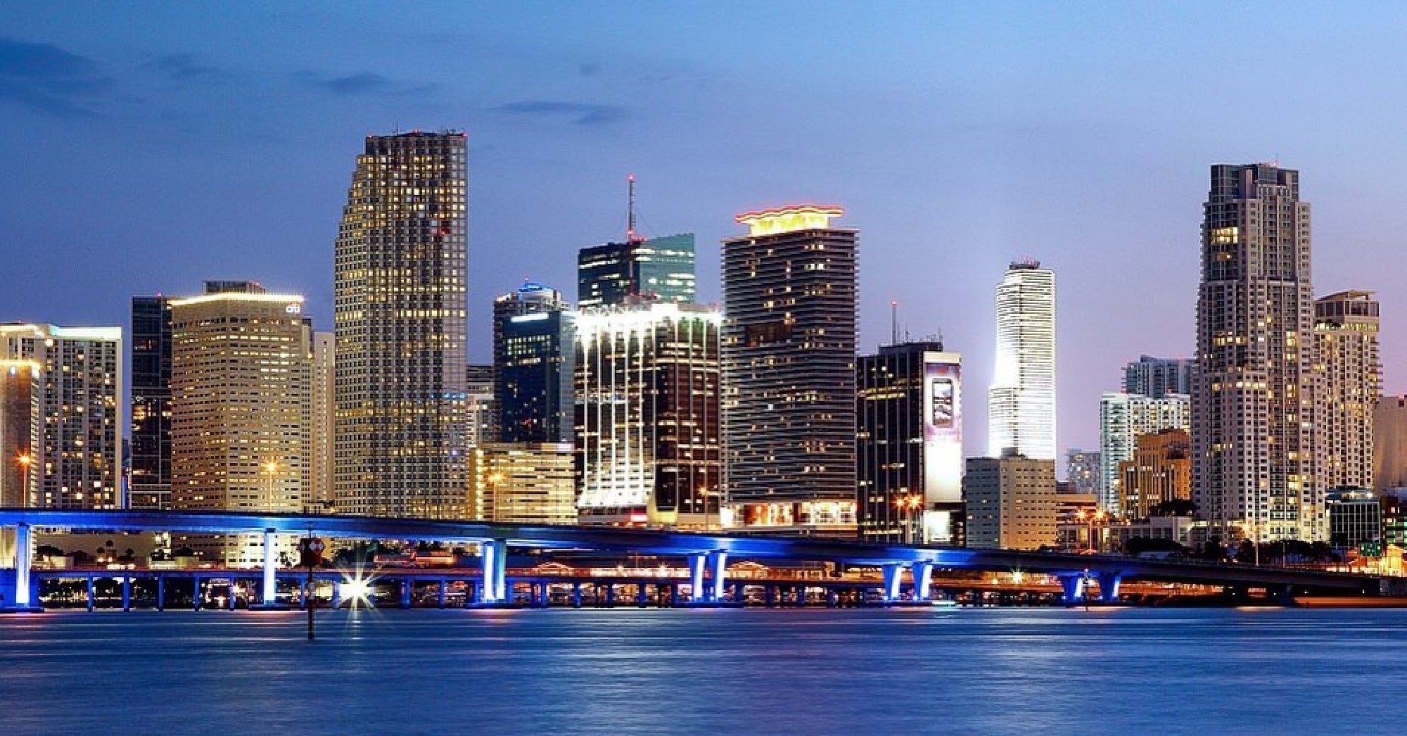 Skyline of a city at dusk with illuminated skyscrapers and a lit bridge over the water in the foreground.