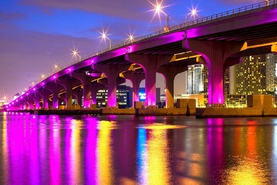 A bridge illuminated with purple and yellow lights spans over water with reflections. City skyline in the background at dusk.