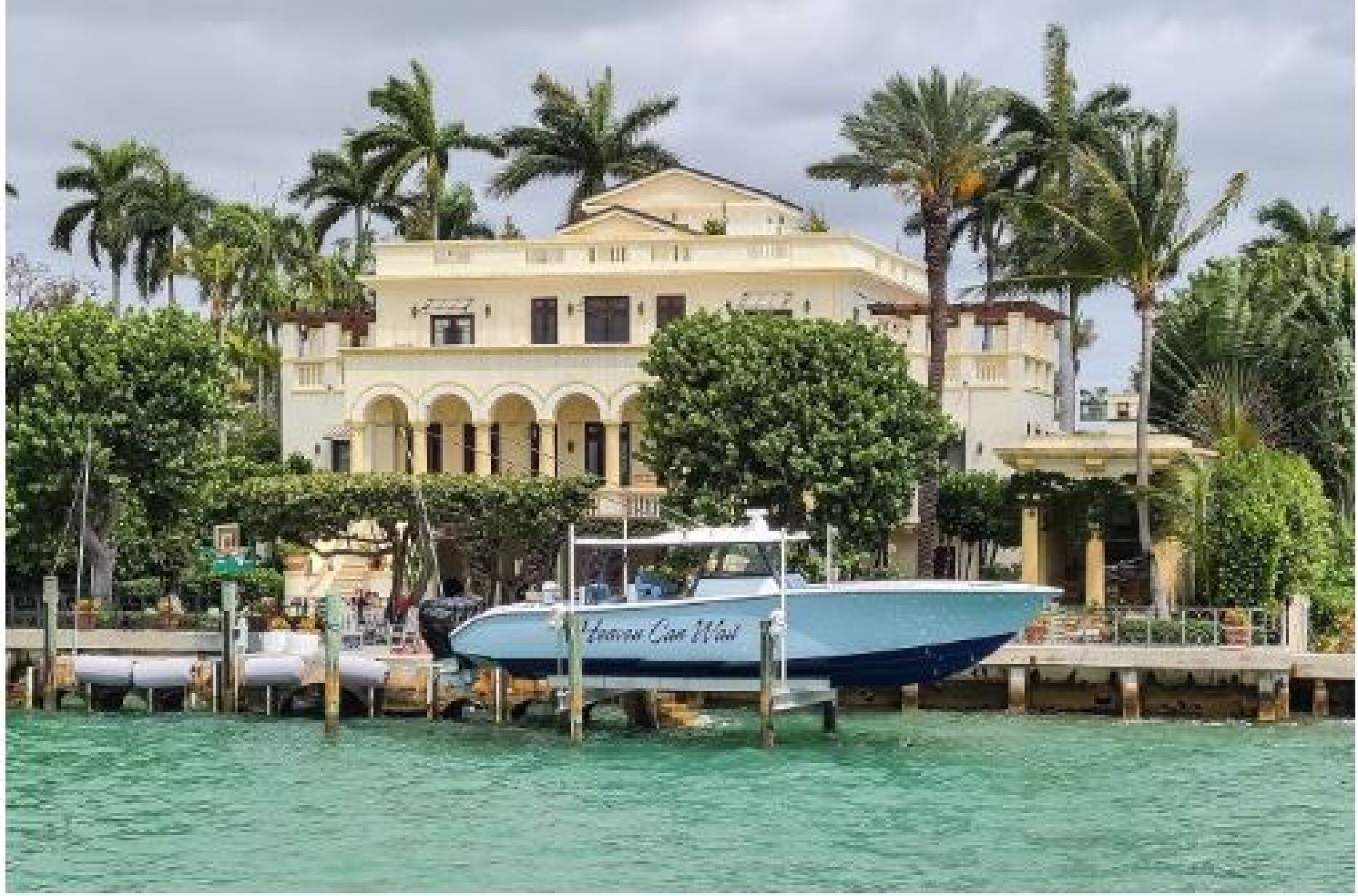 A luxury waterfront villa with a docked white and blue boat, surrounded by palm trees and lush greenery.