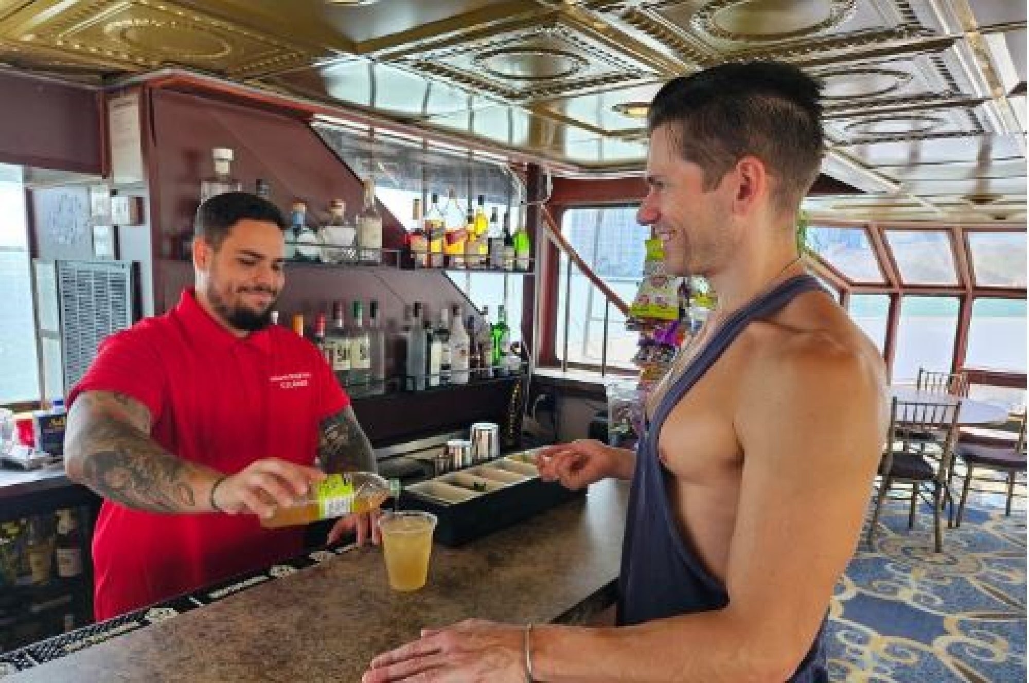 Bartender in a red shirt pours a drink for a smiling man at a bar counter on a boat with windows showcasing a water view.