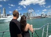 A man and woman stand on a boat looking towards a city skyline with a Ferris wheel. Clouds are scattered across the blue sky.