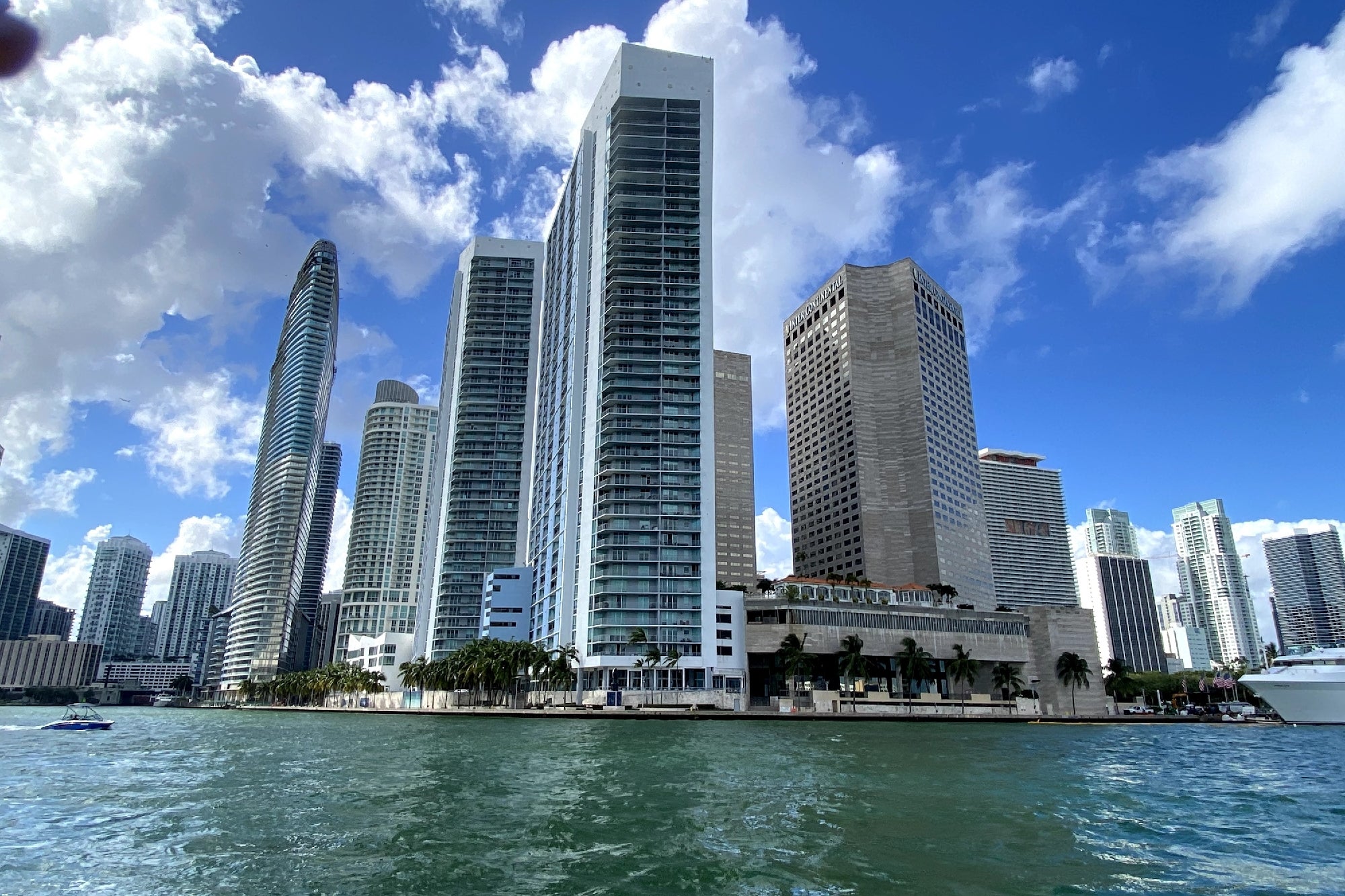 Tall modern skyscrapers line the waterfront under a partly cloudy sky, with water and boats visible in the foreground.