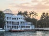 Modern white waterfront mansion with large windows, multiple balconies, and a glass dome roof, surrounded by palm trees at sunset.