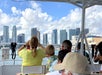 People sit on a boat looking at a city skyline with tall buildings under a partly cloudy sky; some passengers are taking photos.