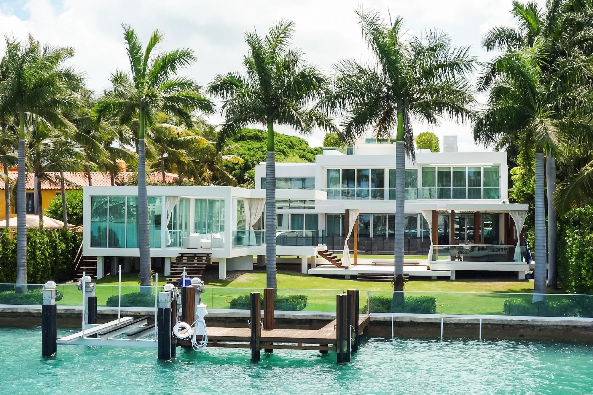 Modern waterfront house with large windows, surrounded by palm trees, featuring a private dock and a lawn, with clear blue water in the foreground.