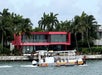 A water taxi boat passes by a modern red and glass house on a waterfront lined with palm trees.
