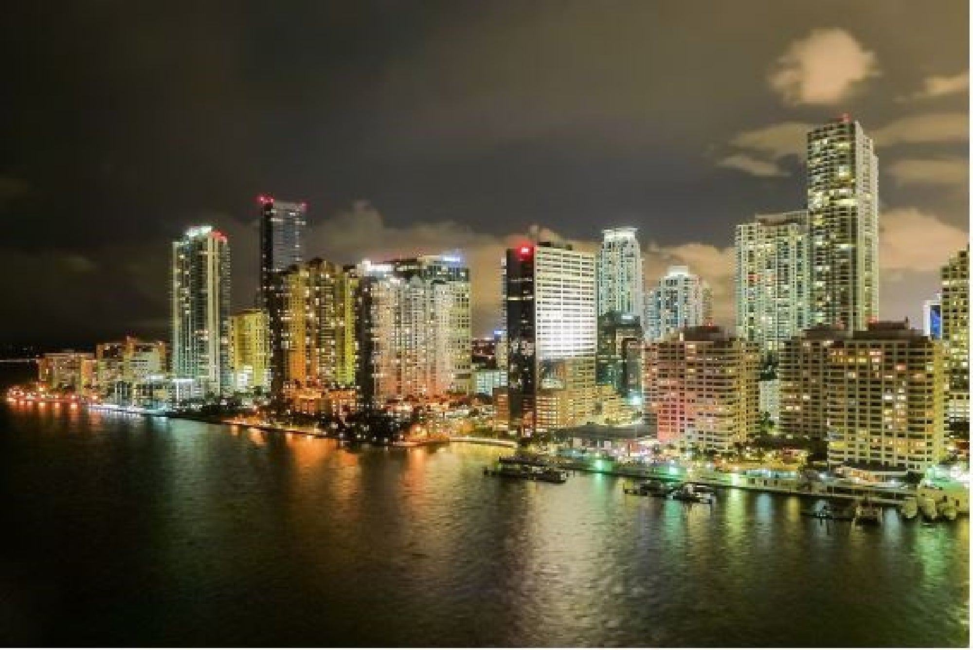 A city skyline at night; tall, illuminated buildings reflect on the calm water below, under a partly cloudy sky.