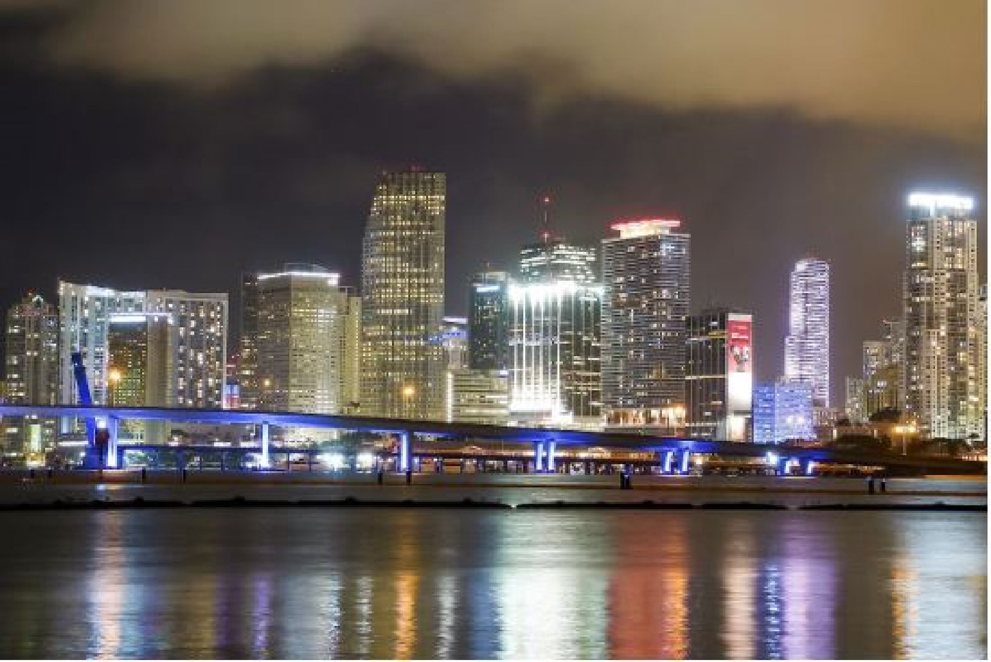 City skyline at night with illuminated buildings and cloudy sky, reflecting on a calm body of water in the foreground.