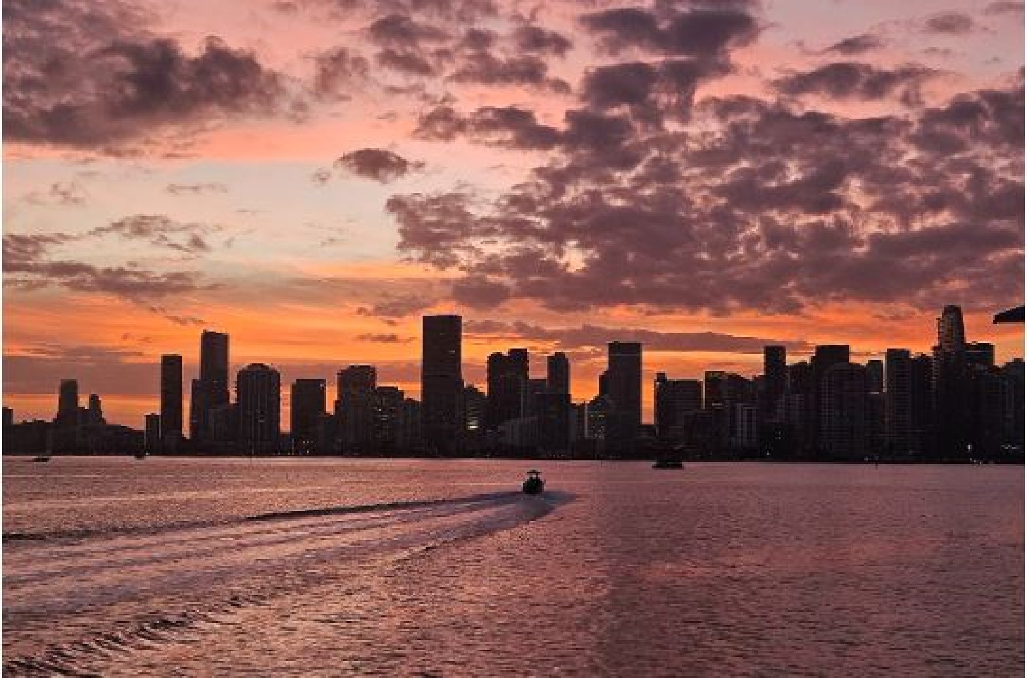 A small boat moves across water toward a city skyline silhouetted against a vibrant sunset sky with scattered clouds.
